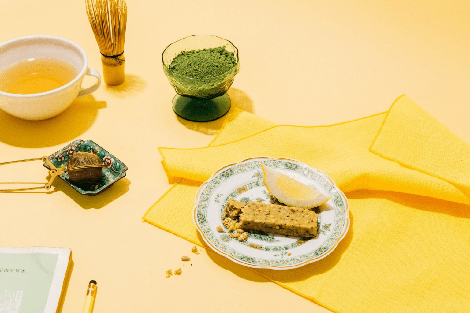 Dang Foods lemon snack bar sits on a plate surrounded by yellow elements such as tablecloths and tea. There is also matcha and a matcha whisk next to this organic snack.