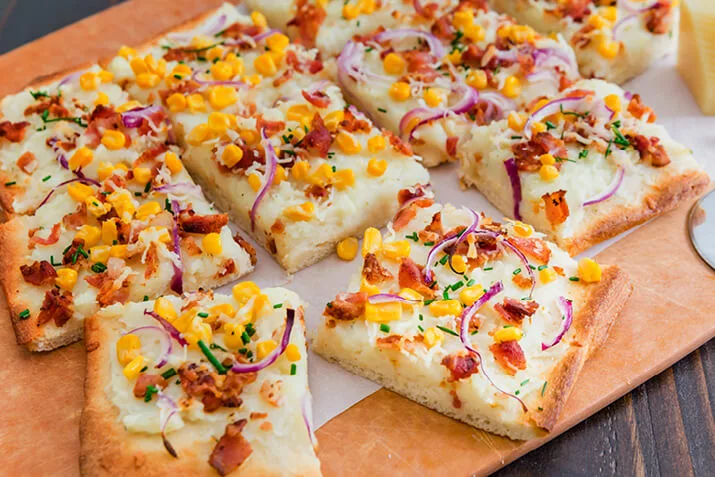 A mashed potato pizza sits on a cutting board, cut into squares, ready to be eaten.