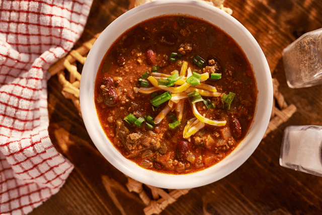A bowl of chili sits next to salt and pepper and is topped with cheese and onion.
