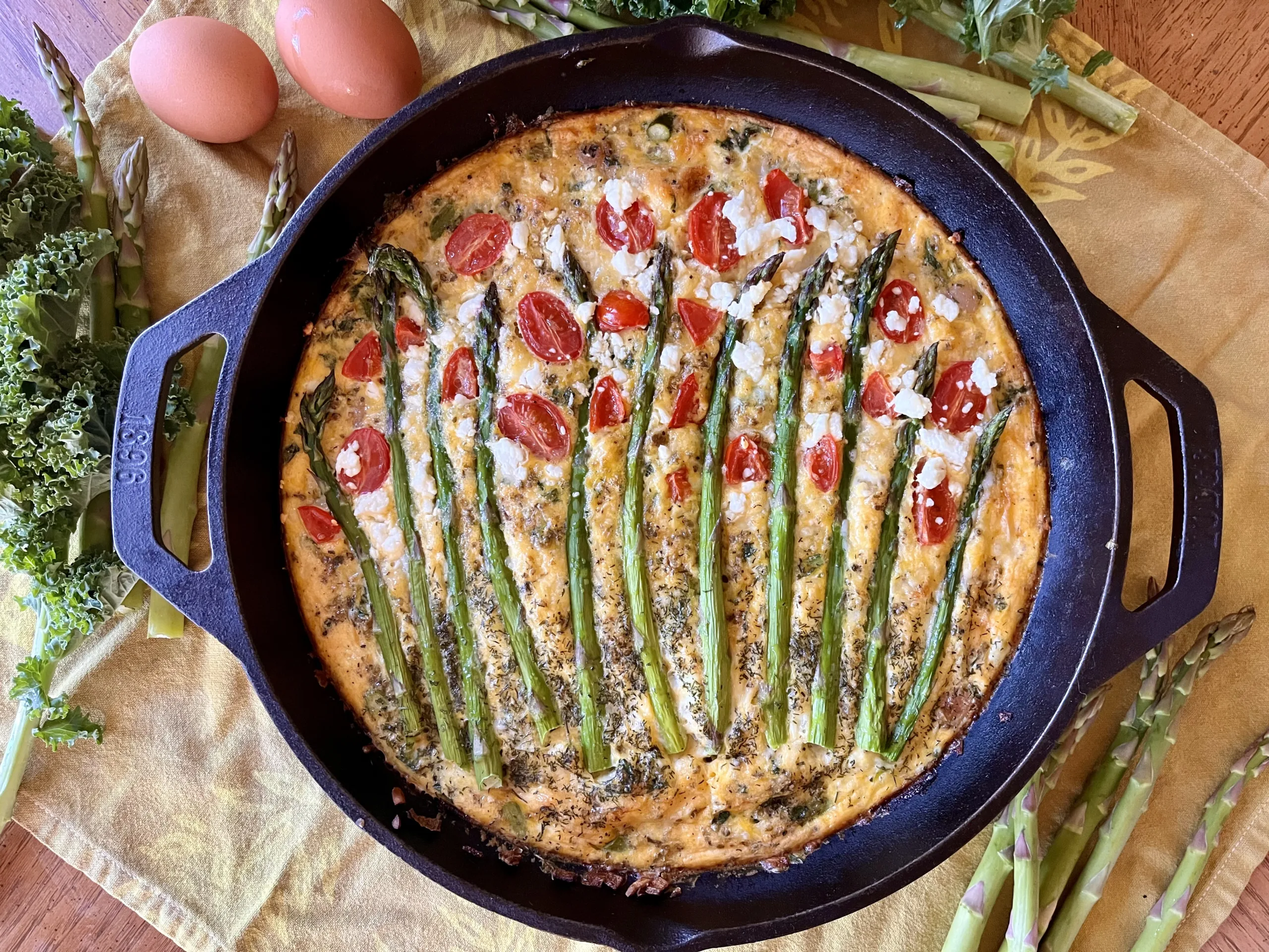 A finished frittata sits on a counter, ready to be enjoyed. It is topped with asparagus and tomatoes.