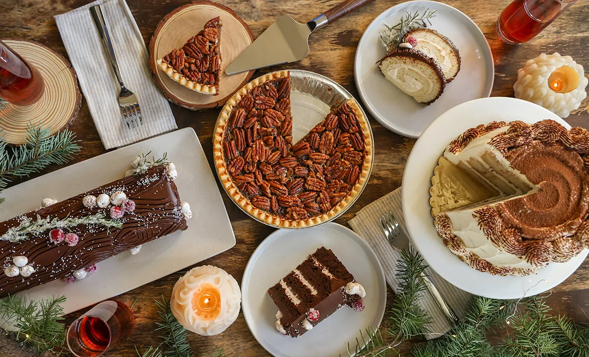 a table full of holiday desserts including buche de noel cake, pecan pie, and tiramisu cake 